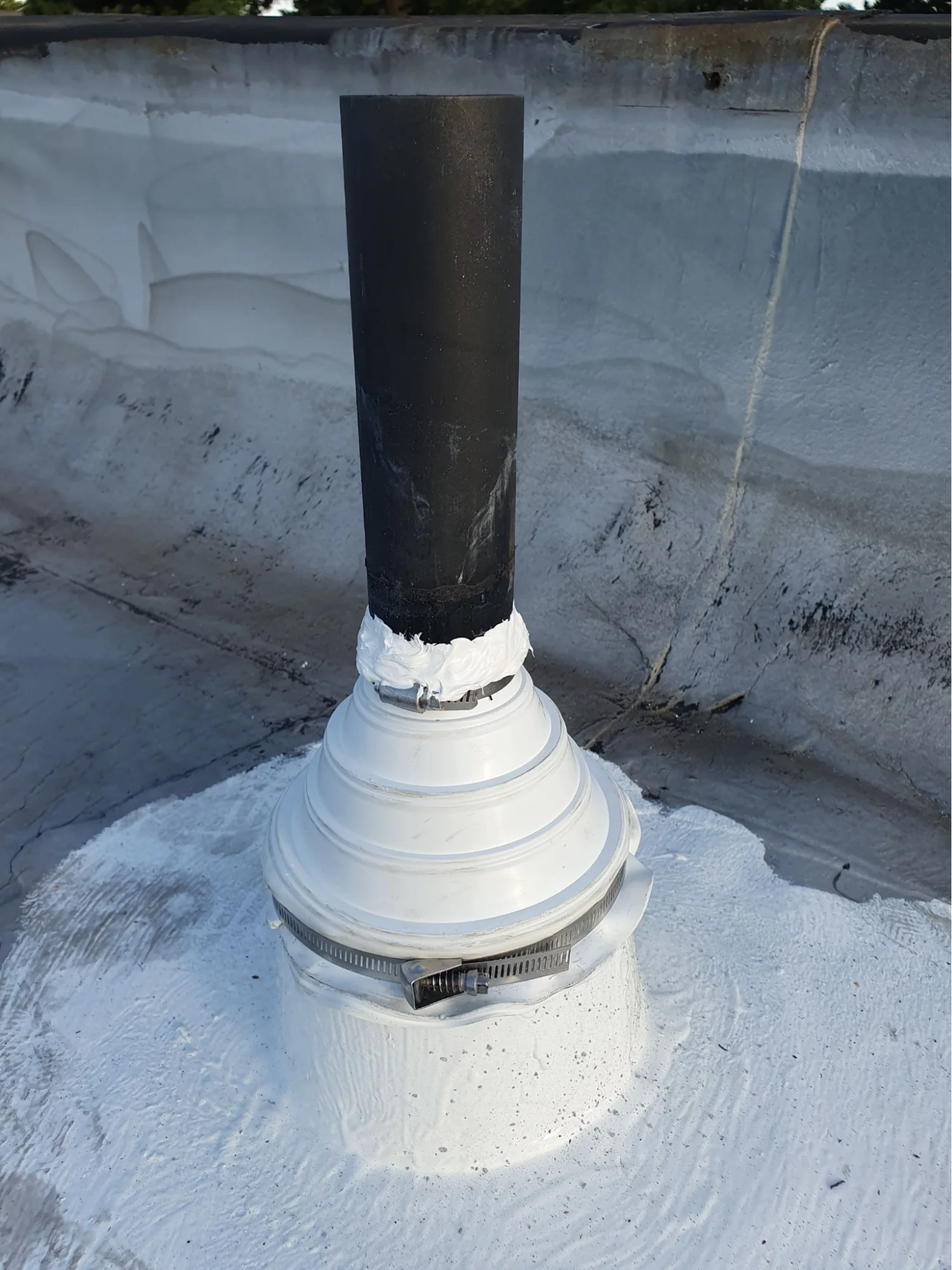 A black vent pipe on a white silicone-coated flat roof, fitted with a white pipe boot clamped at the base and sealed into the surface, with a coated parapet wall behind.
