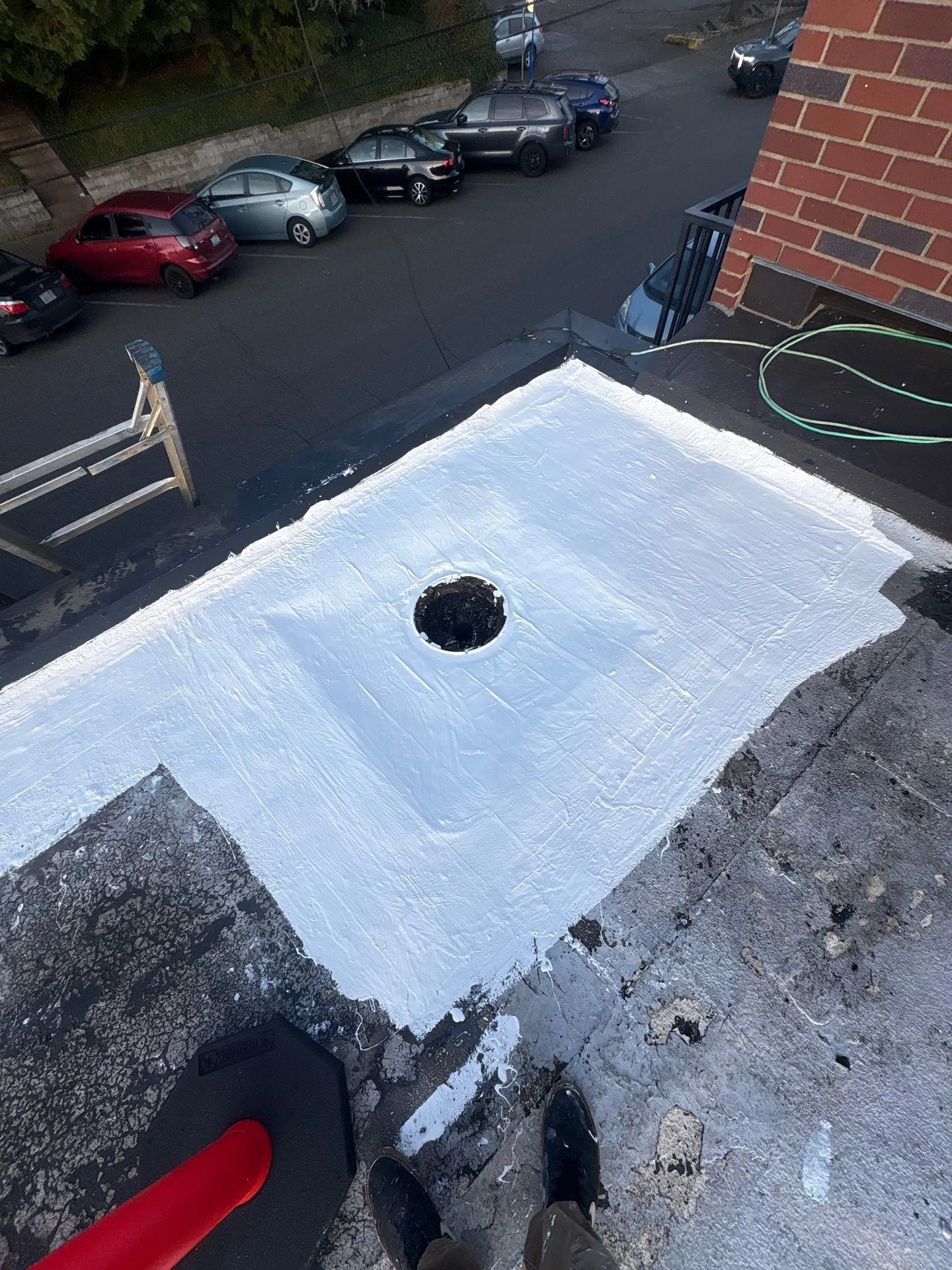 An overhead view of a flat roof drain freshly coated in white silicone around its opening, the sealed white field standing out against the dark surrounding roof, with a parking lot of cars below.
