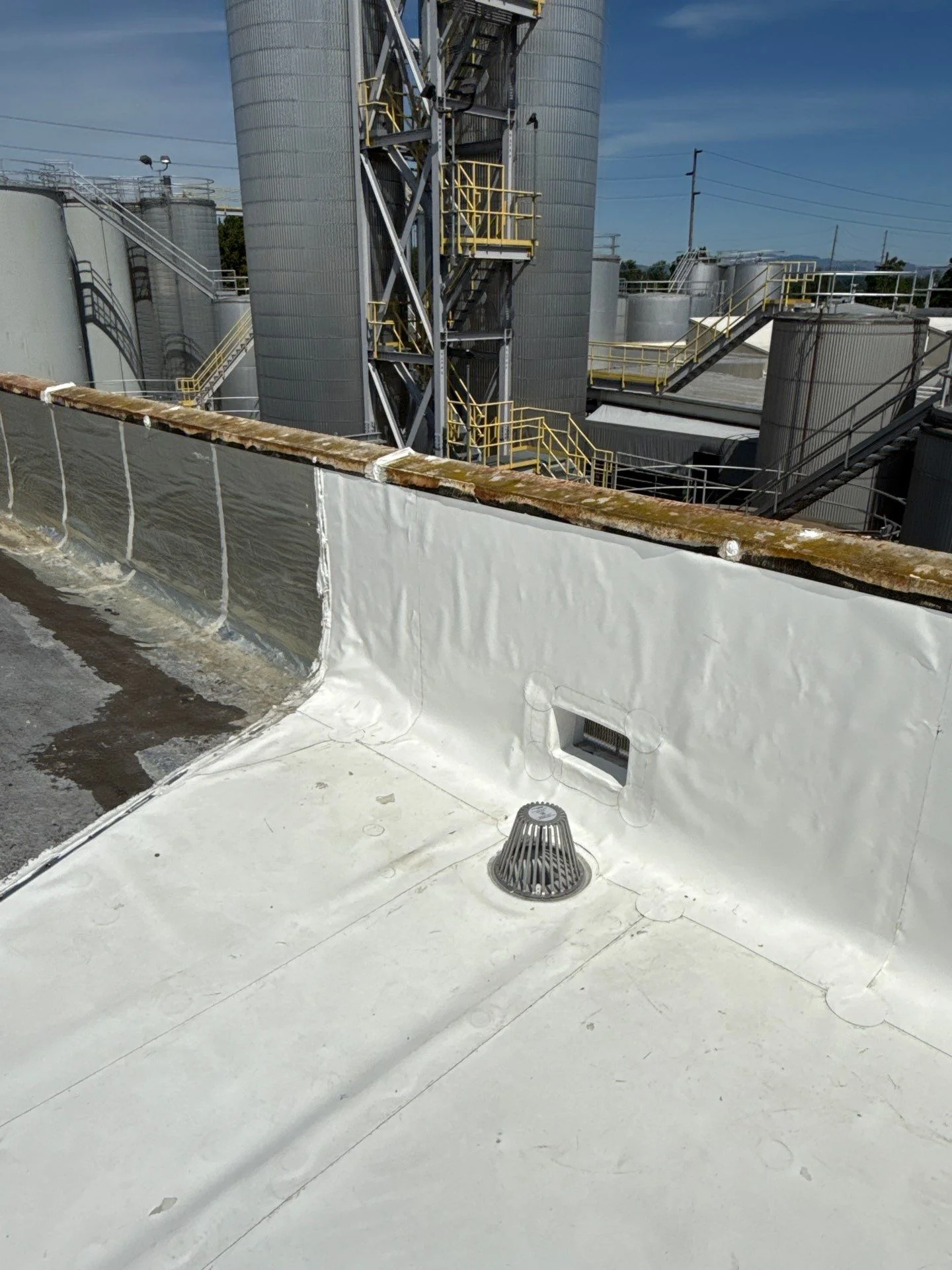 A white flat roof on an industrial facility, membrane wrapped up the parapet and welded around the drain and scupper, with steel storage silos, walkways, and yellow safety railings rising in the background.