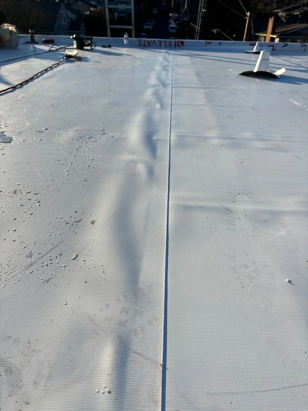 A heat-welded seam running down a bright white flat-roof membrane in sunlight, with the Elevate brand stripe along the parapet and a vent boot at the upper edge.