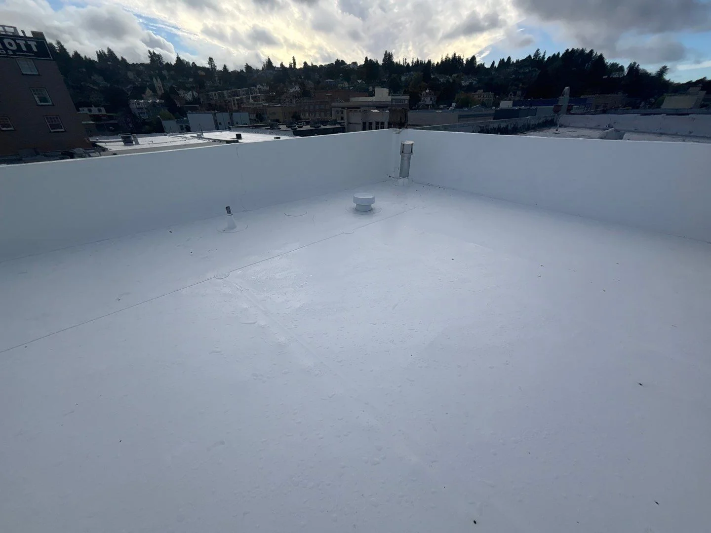 A fresh white flat-roof membrane after rain, beads and shallow pools of water sitting on top of the surface, white parapet walls wrapping the perimeter, with a hillside city view under a breaking cloudy sky beyond.
