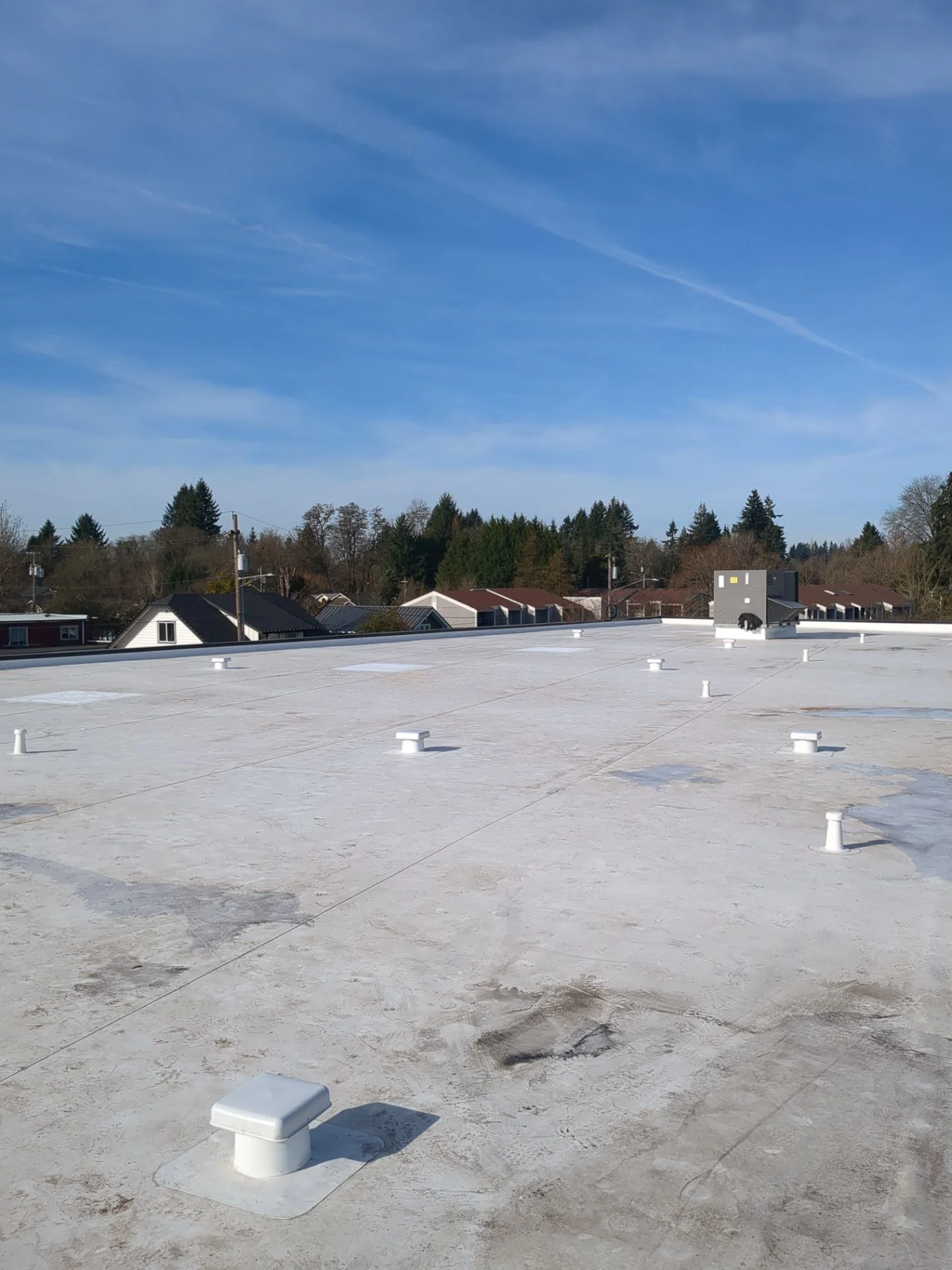 A wide white flat-roof membrane stretching to a tree-lined horizon, vents and an HVAC unit set across its surface, with visible foot traffic and weathering marks on the wear layer.