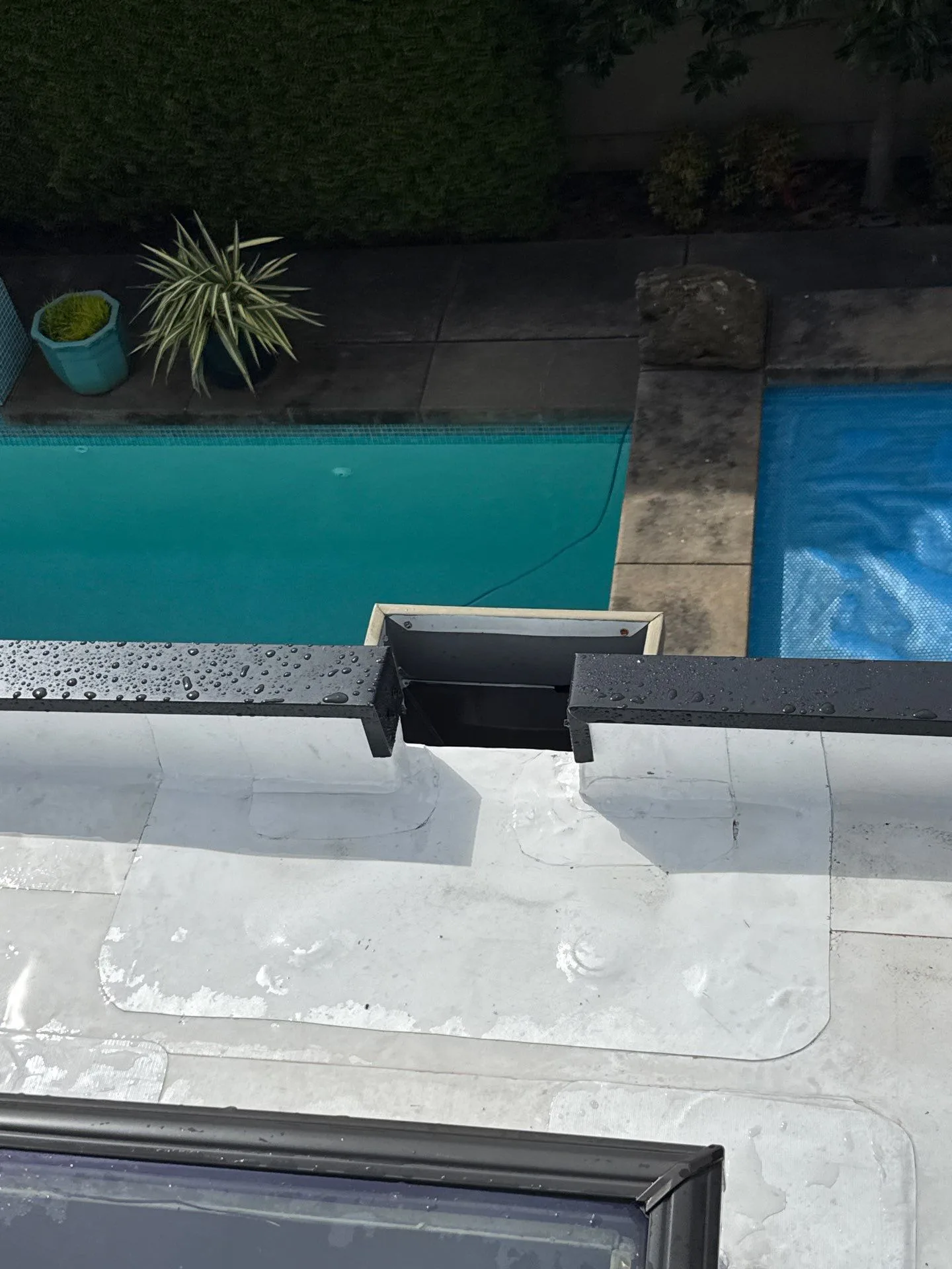 A through-wall scupper cut into a white membrane parapet wall, the membrane wrapped and welded into the opening, with a swimming pool visible on the ground below.