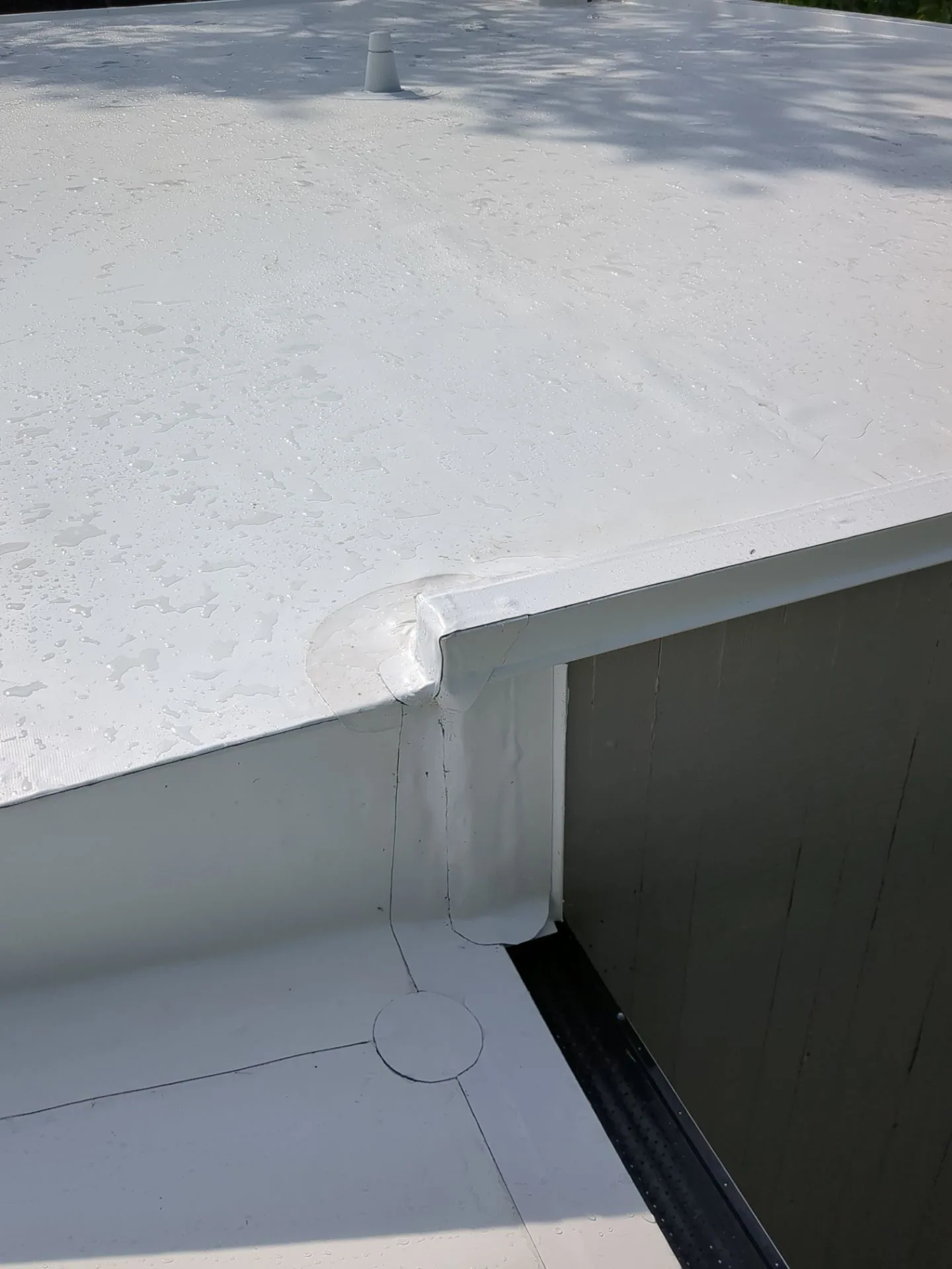 A fresh white flat-roof membrane sloping gently toward a finished metal drip edge, beads of rainwater running off the surface, with a vent boot in the background.