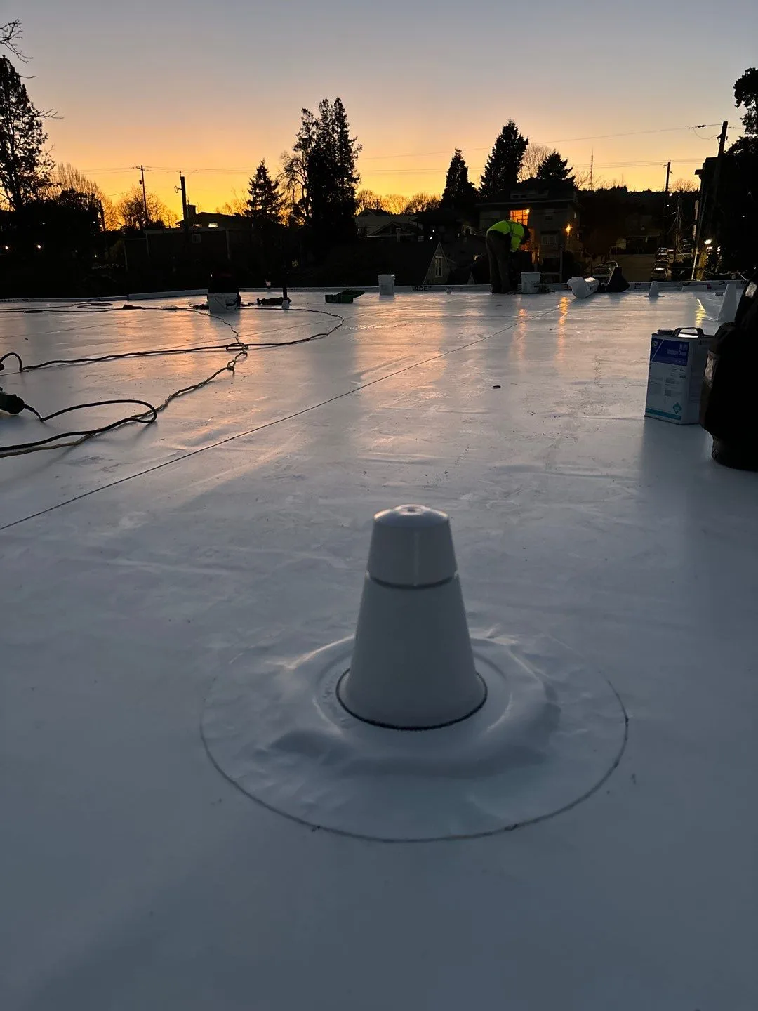 A white vent boot heat-welded into a fresh white flat-roof membrane, its base sealed in a clean ring, with the roof stretching out under an orange sunset sky.