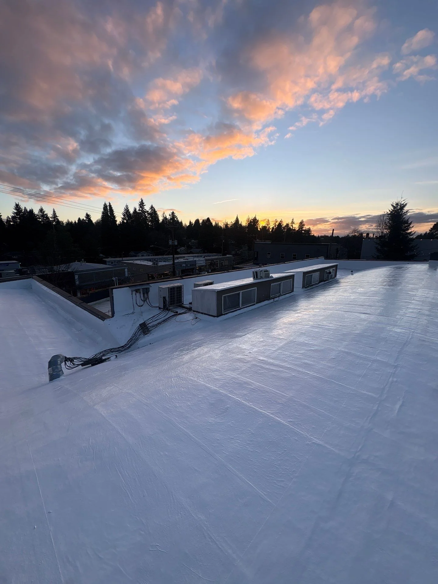A white-coated flat commercial roof stretching toward a vivid sunset, its smooth sealed surface holding up beside rooftop units and a parapet as the sky turns orange over a distant tree line.