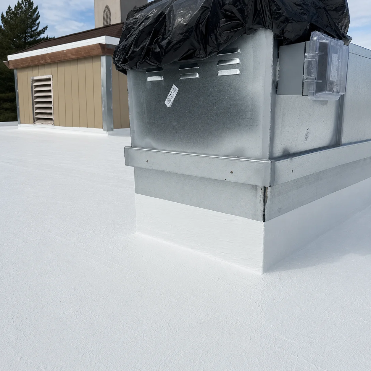 A corner of a TPO flat roof freshly coated in bright white silicone under a blue sky, the coating carried up the metal curb of a tarp-covered rooftop unit, with a tan outbuilding and evergreens behind it.