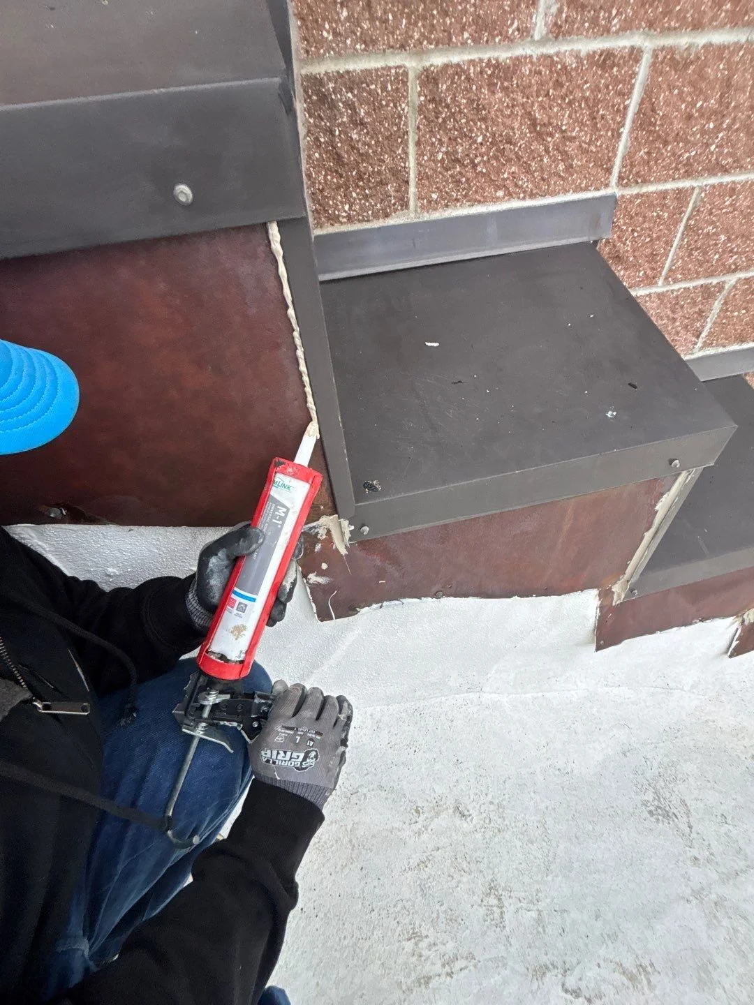 A gloved worker running a bead of sealant along the metal flashing where a flat roof meets a brick wall and a raised metal-capped curb, tying the wall and edge details into the sealed surface.
