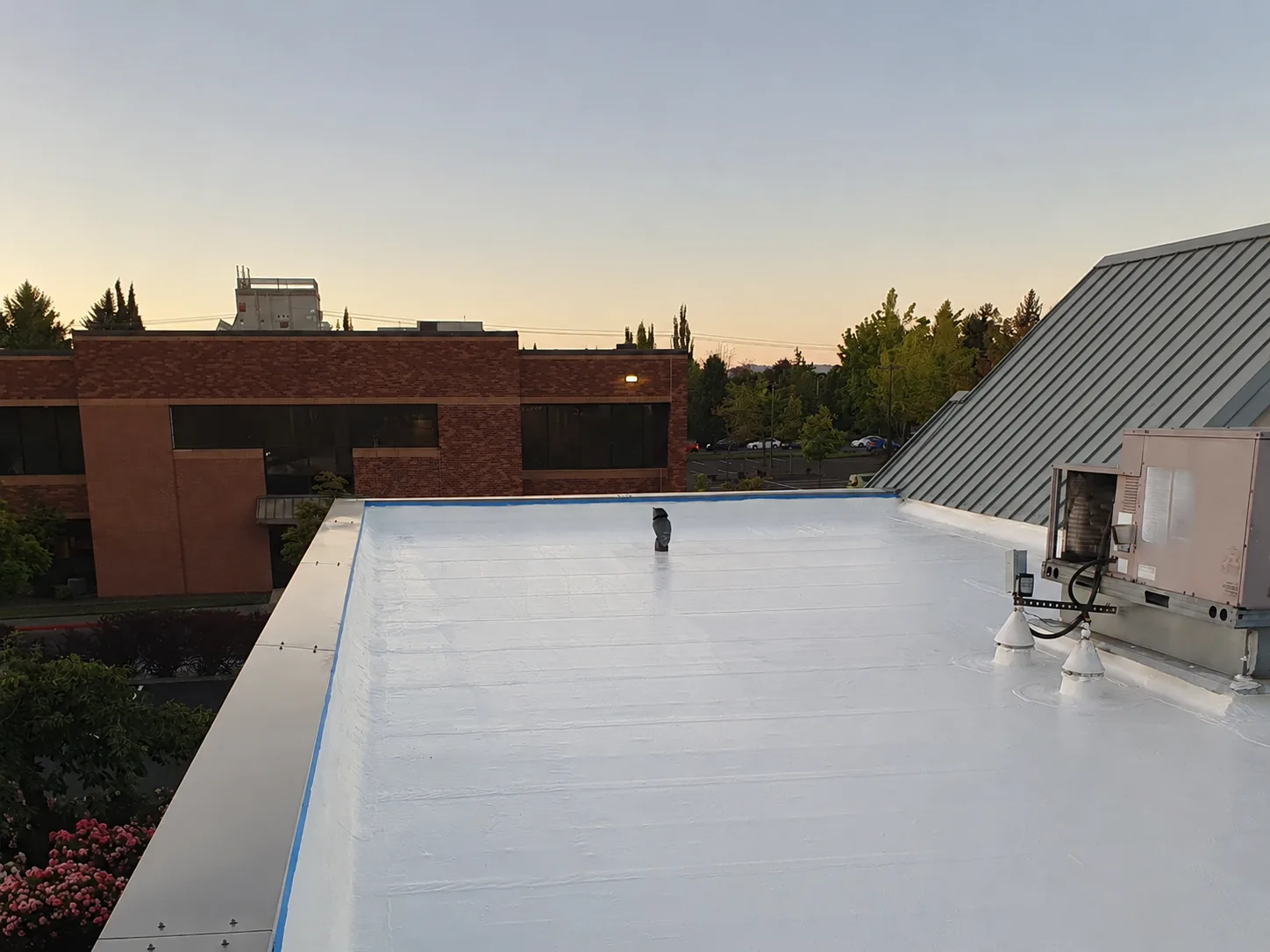 A freshly renewed white flat roof on a commercial building at dusk, finished up to its parapet beside a brick wing and metal mansard.