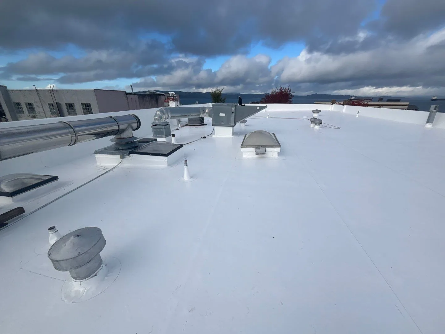 A freshly installed white flat roof with vents, ducting, and skylights sealed in, overlooking water under a dramatic cloudy sky.
