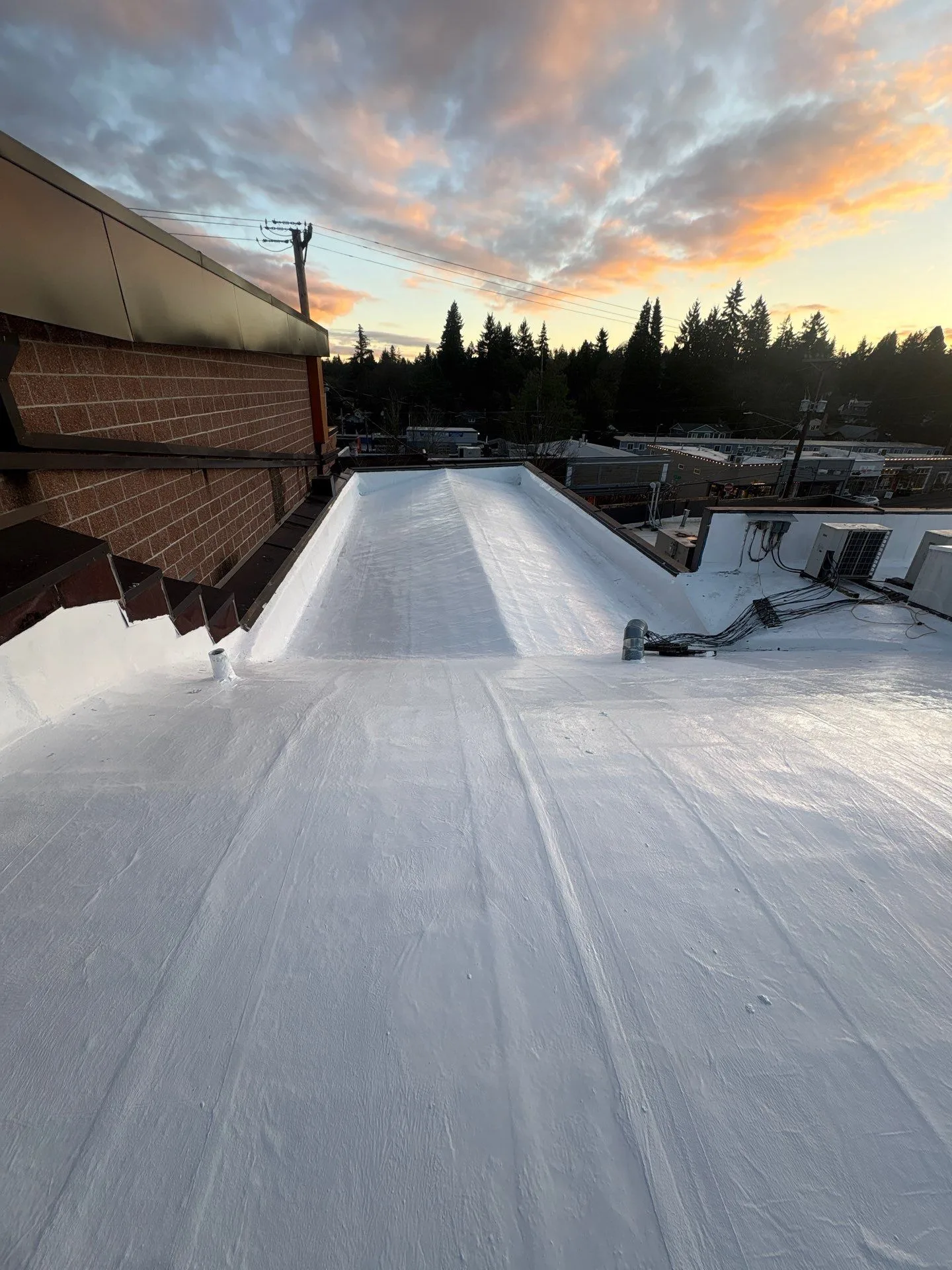A flat commercial roof freshly renewed with a seamless white silicone coating, running between brick parapet walls toward a town and tree line under a sunset sky.