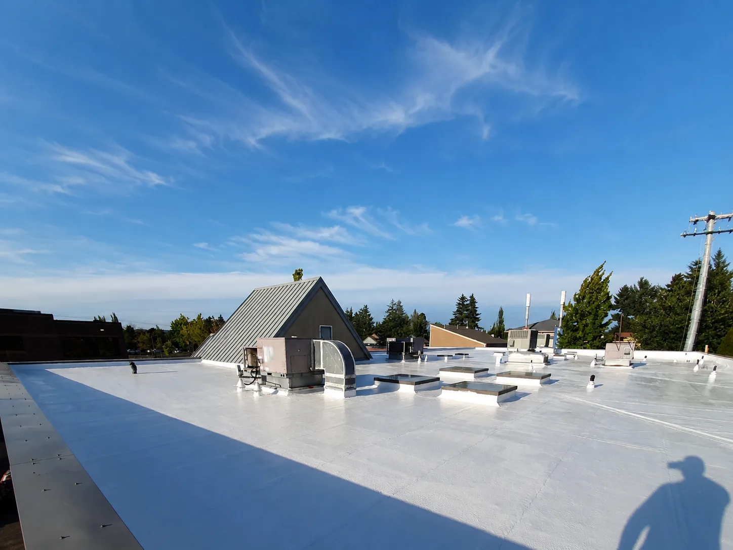 A wide flat commercial roof renewed with one seamless white silicone surface stretching around an A-frame dormer, vents, and skylights under a bright blue sky.